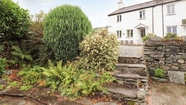A white building with a stone staircase leading to a door, surrounded by lush greenery and a stone wall. - Hilber Cottage (Ambleside)