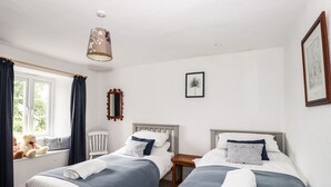 A bedroom with two beds, a window with curtains, a bedside table, a mirror, and a hanging light fixture. - Hilber Cottage (Ambleside)