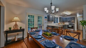 A dining area with a wooden table set for six, surrounded by chairs. A console table with a lamp and decorative items is to the left. The room has a chandelier, a view through the door, and a kitchen in the background. - Spacious Mountain Home with Fireplace, Game Room in Bear Creek Lake Community! (Jim Thorpe)