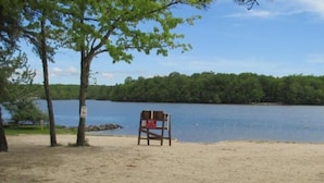 A beach chair with a 'No Swimming' sign, a sandy beach, and a lake with trees in the background. - Spacious Mountain Home with Fireplace, Game Room in Bear Creek Lake Community! (Jim Thorpe)