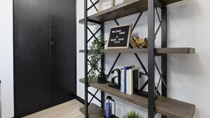 Una habitación con un armario negro, una banca de madera y una estantería metálica con diversos objetos, incluyendo libros, una planta pequeña y un cartel de bienvenida.