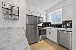 A modern kitchen with white cabinets, stainless steel appliances, a marble countertop, and a window with blinds.
