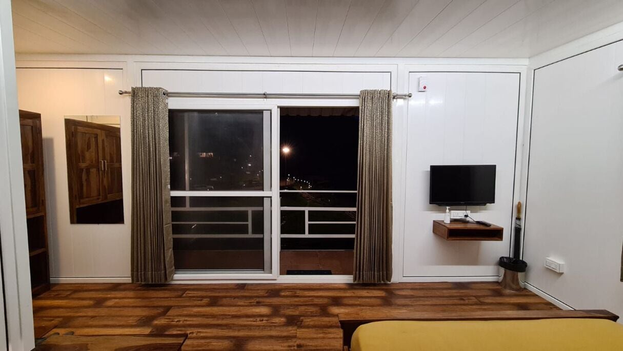 A room with a wooden floor, a bed, a TV on a shelf, and a sliding glass door leading to a balcony.