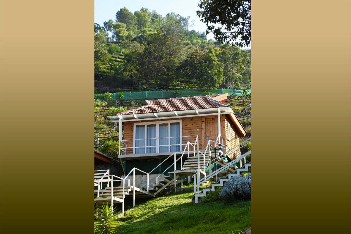 A wooden cabin with a tiled roof, surrounded by lush greenery and a hillside.