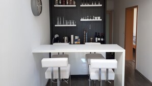 Et moderne køkken med et hvidt barområde, to hvide barstole og reoler med diverse køkkenartikler og flasker.