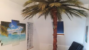 En stue med et stort, kunstigt palmetræ, en sort lædersofa og et lille træbord.