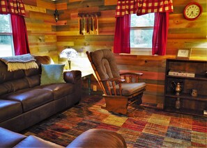 Ein gemütliches Blockhaus-Interieur mit einem Ledersofa, einem Holzsesseln, einem gemusterten Teppich, einer Lampe und einem Regal mit einer Kaffeemaschine.