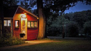 Eine kleine, rote Holzhütte mit beleuchtetem Eingang, umgeben von Bäumen und einer Schaukel in einem Baum.