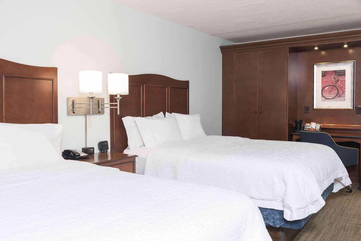 A hotel room with two beds, a nightstand with a phone and a lamp, a desk with a chair, and a framed picture on the wall.