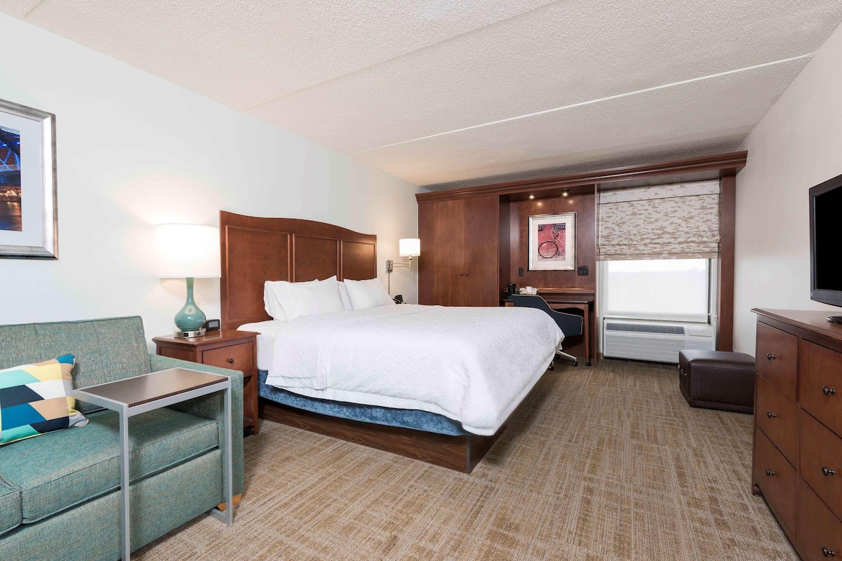 A hotel room with a bed, a desk, a chair, a dresser, a TV, a window with a view, and a framed picture on the wall.