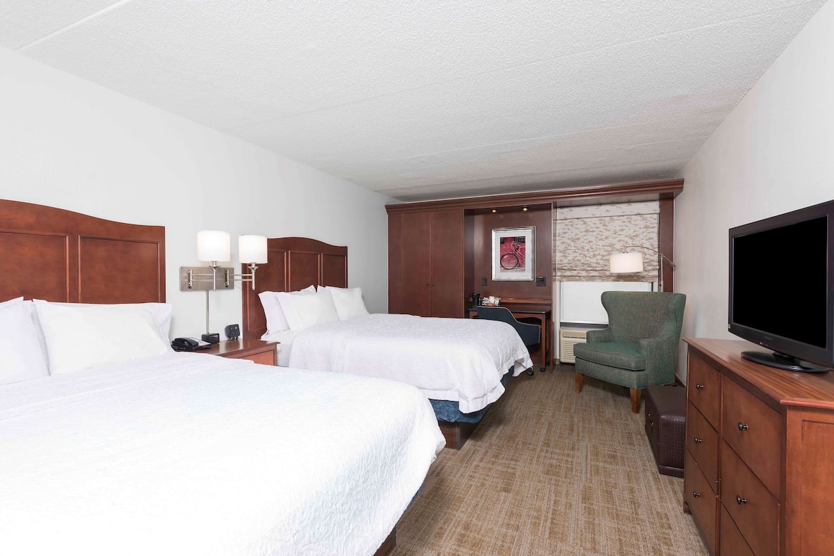 A hotel room with two beds, a television, a desk with a lamp, and a chair.