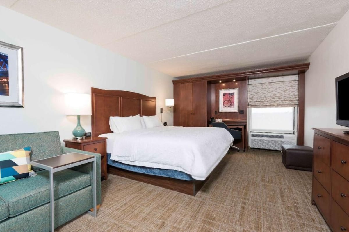 A hotel room with a bed, a desk, a chair, a TV, a window, and a dresser.