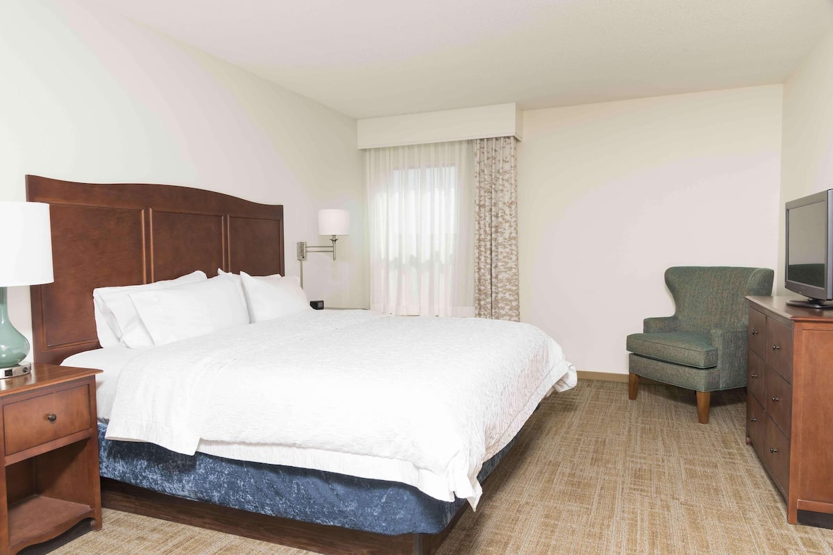 A hotel room with a wooden bed, a green armchair, a TV on a wooden cabinet, and a nightstand with a lamp.