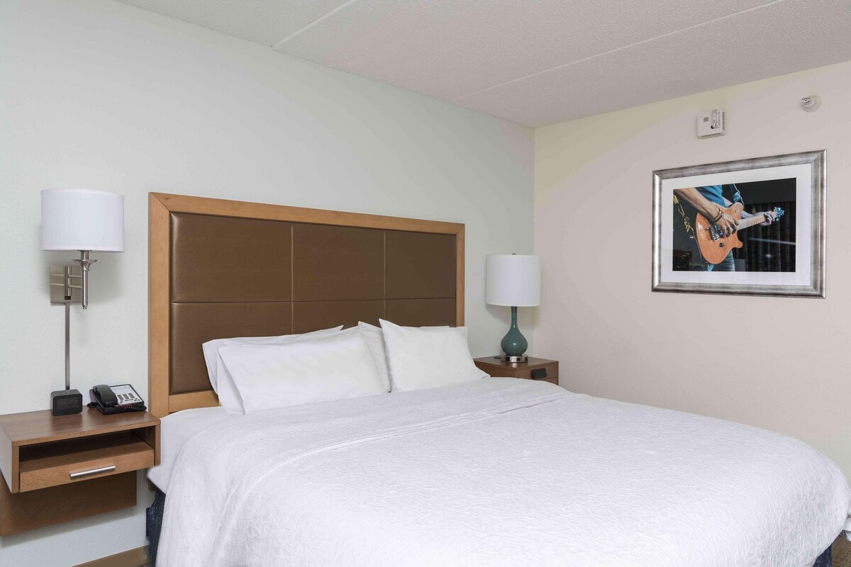 Une chambre d’hôtel avec un grand lit, deux tables de chevet, une lampe, un téléphone et un tableau encadré au mur.