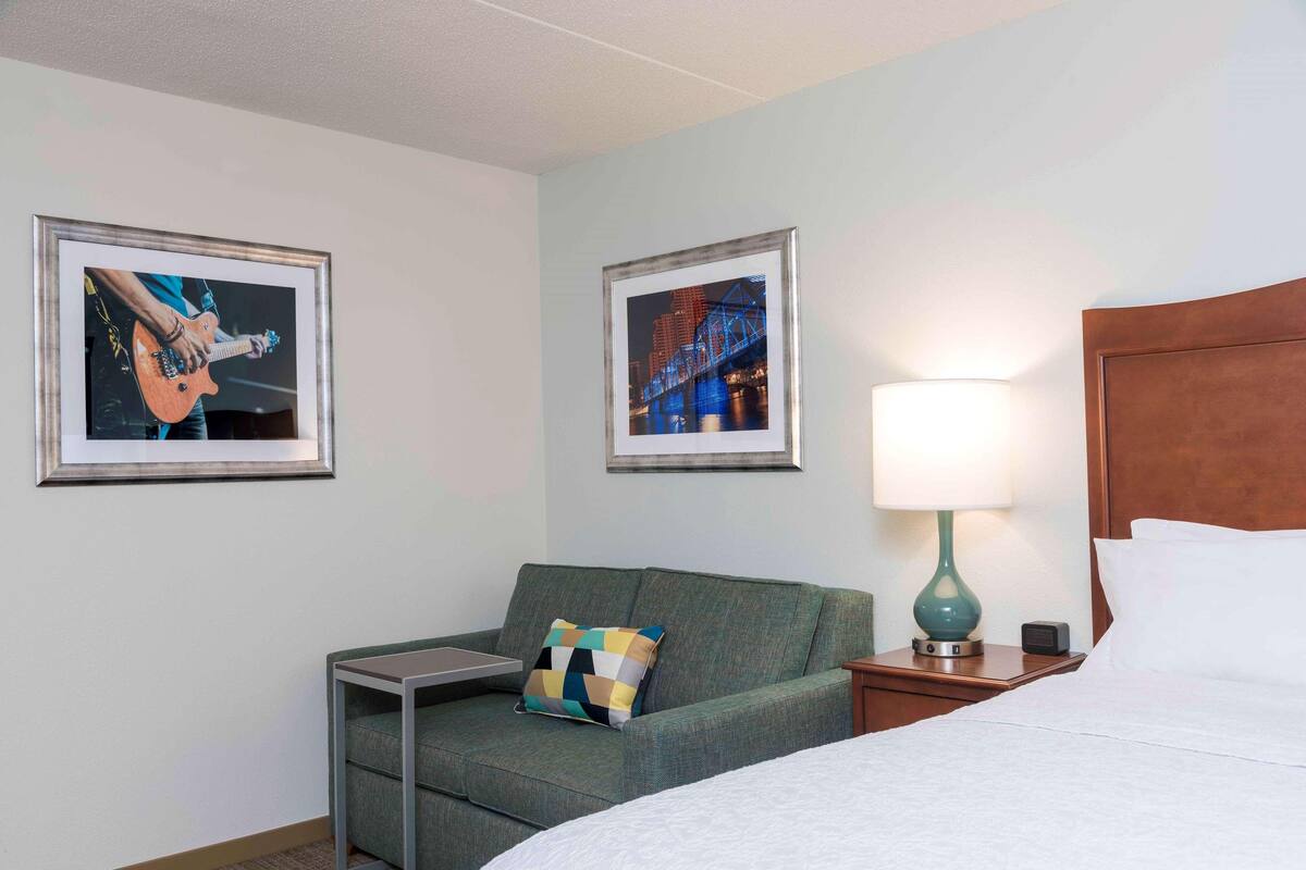A hotel room with a bed, a green sofa, a small table, a lamp, and two framed pictures on the wall.