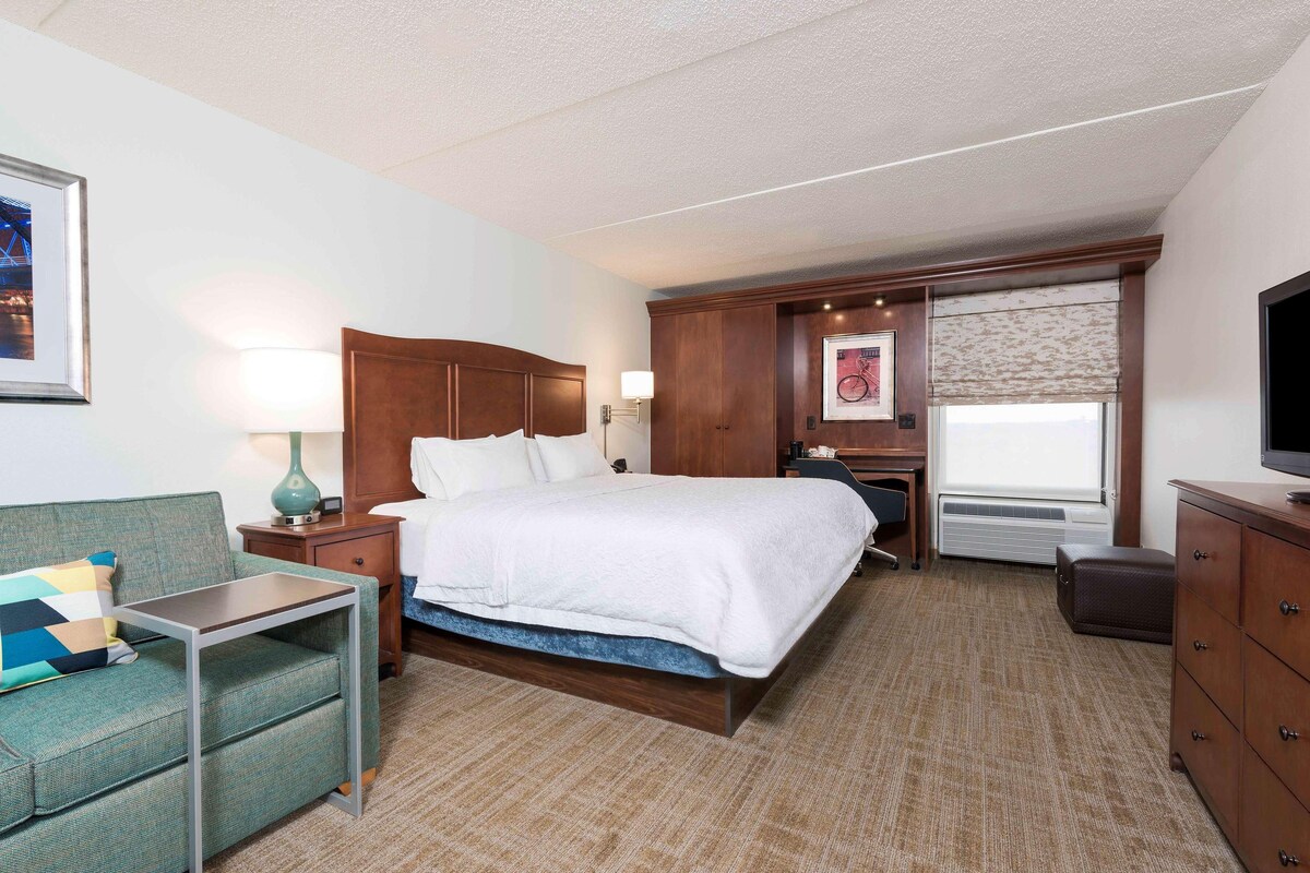 Une chambre d’hôtel comprenant un lit, un bureau, une chaise, une commode, une télévision, une fenêtre avec vue et un tableau encadré au mur.