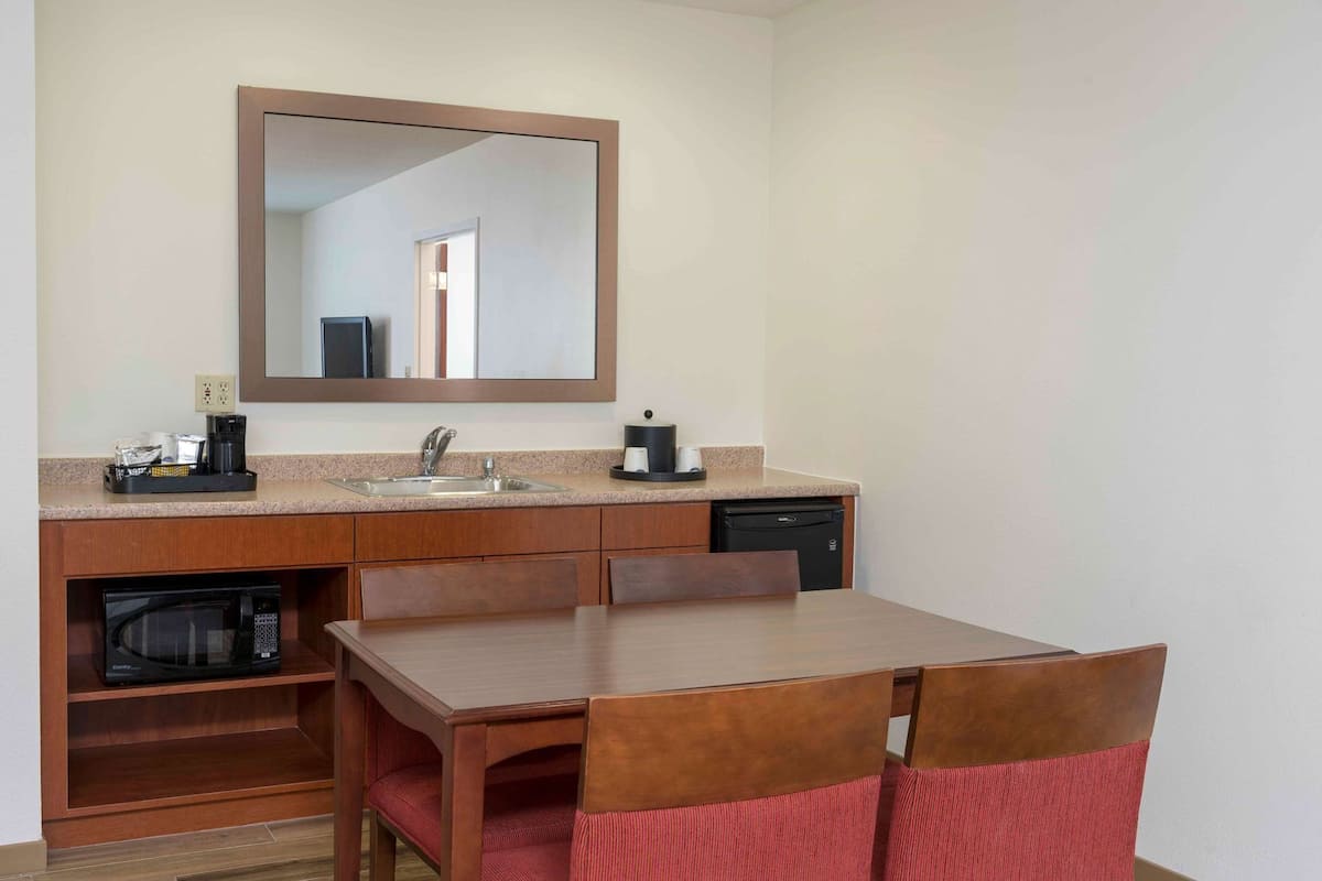 A compact kitchenette with a microwave, sink, and coffee maker, a wooden dining table with chairs, and a large mirror above the counter.