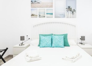 Une chambre à coucher avec un lit blanc, deux tables de chevet, une tête de lit et des œuvres d’art murales représentant des scènes de plage.