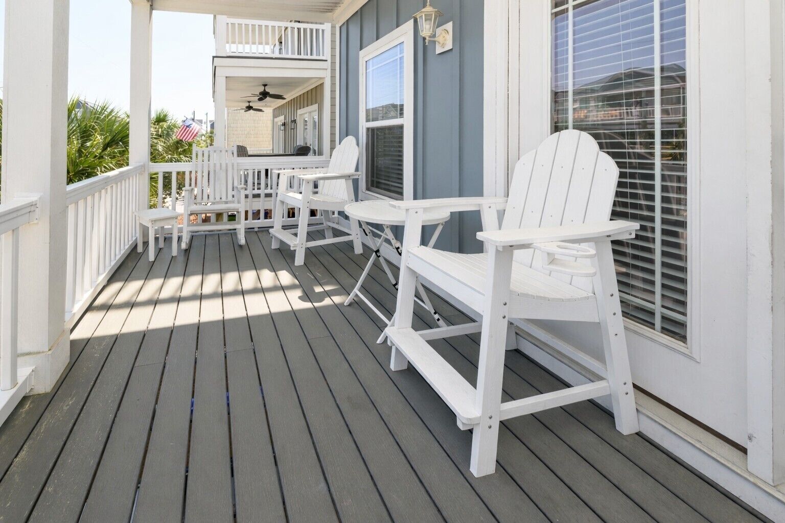 Eine überdachte Veranda mit weißen Adirondack-Stühlen, einem Tisch, einem Deckenventilator und einem Fenster mit Jalousien.