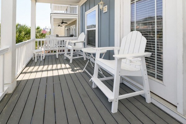 A covered porch with white Adirondack chairs and a table, a ceiling fan, and a window with blinds.