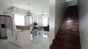 Une cuisine spacieuse dotée d’un plan de travail en marbre, d’un îlot central avec des chaises et d’un escalier en bois sombre menant à un palier.