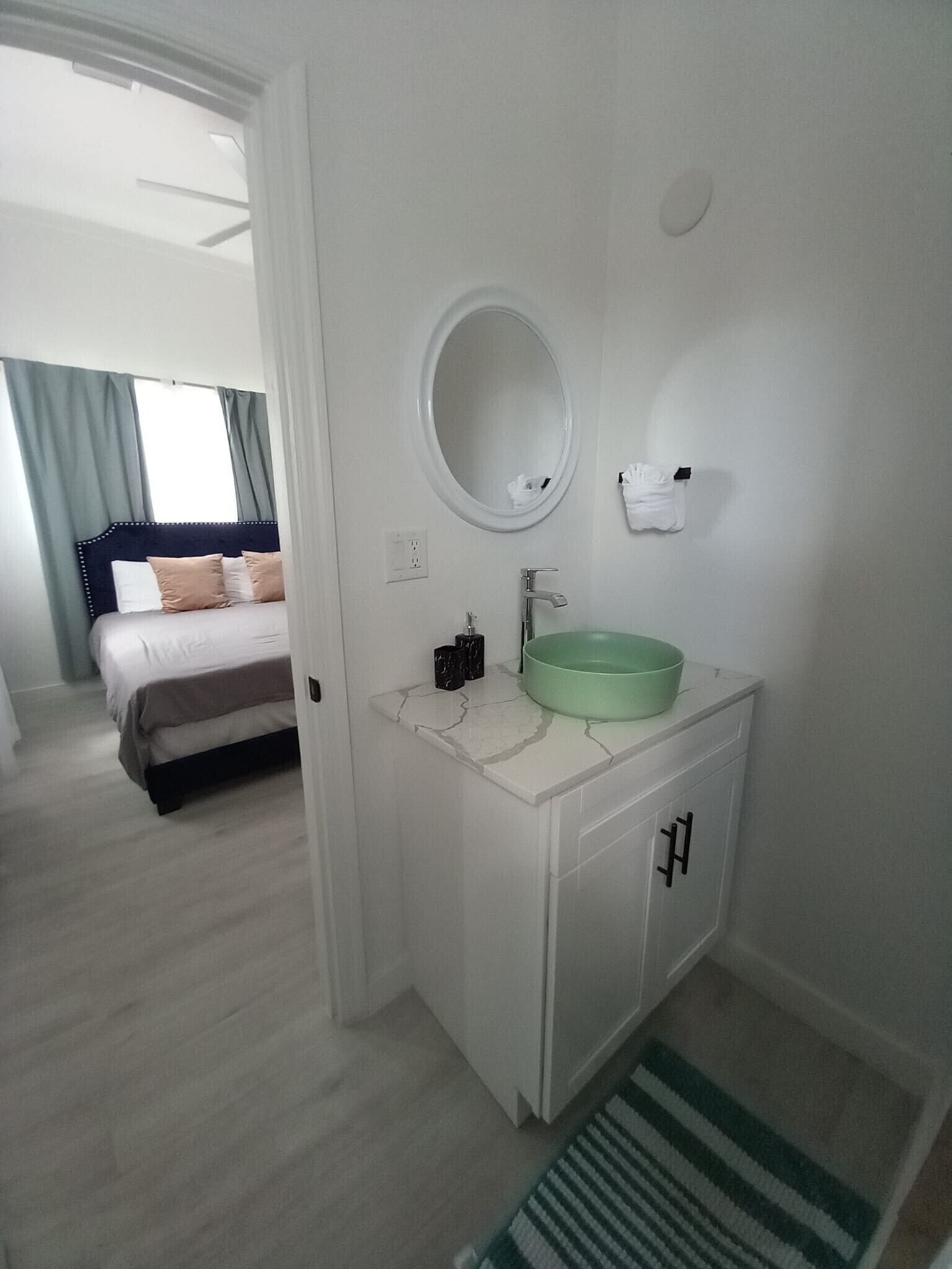 Una habitación de hotel con una cama, un baño con un lavabo verde, un espejo y una alfombra a rayas.