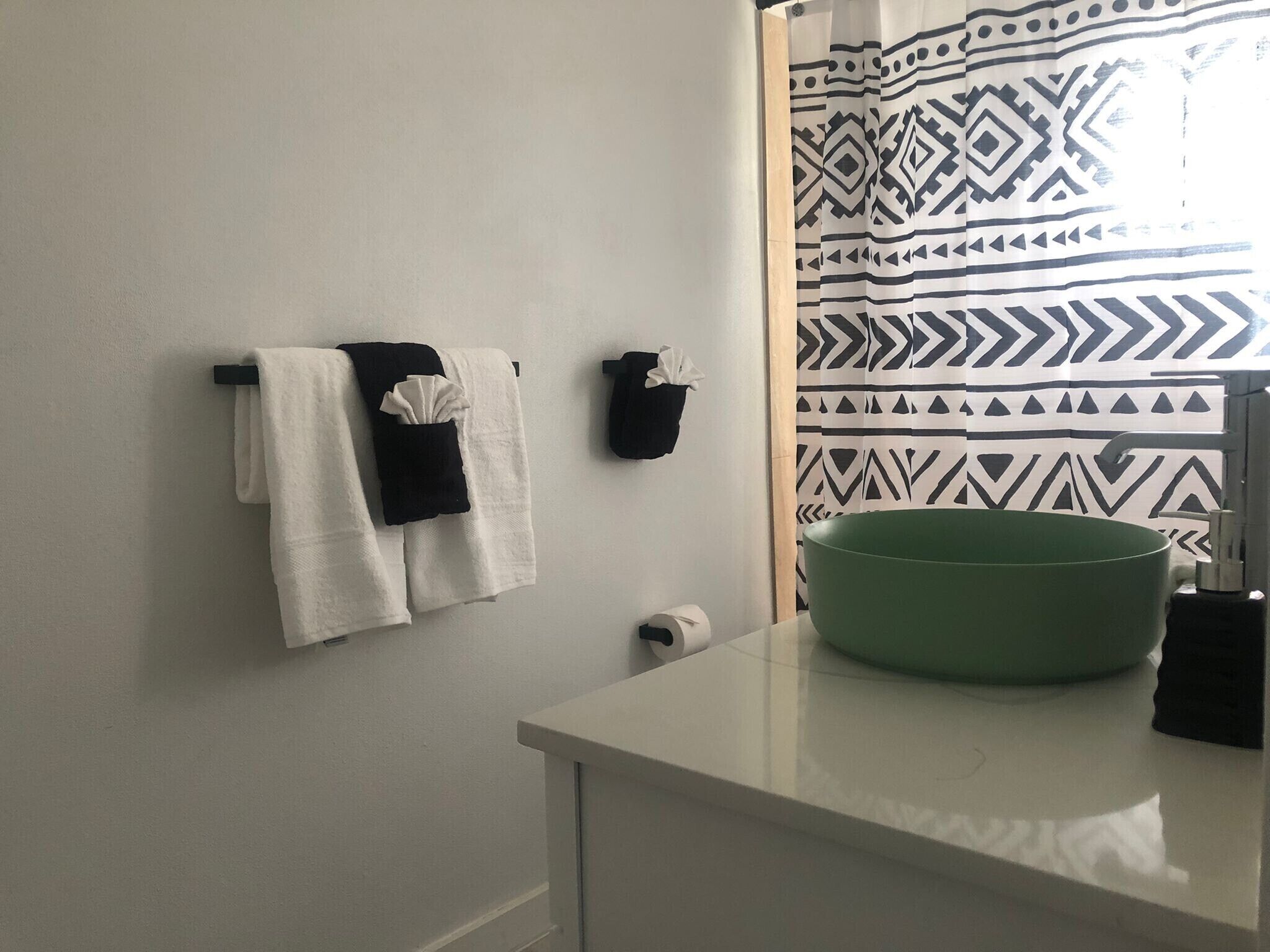 A bathroom with a white sink, a green basin, a towel rack with two towels, a black towel, and a patterned shower curtain.