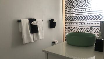 A bathroom with a white sink, a green basin, a towel rack with two towels, a black towel, and a patterned shower curtain.