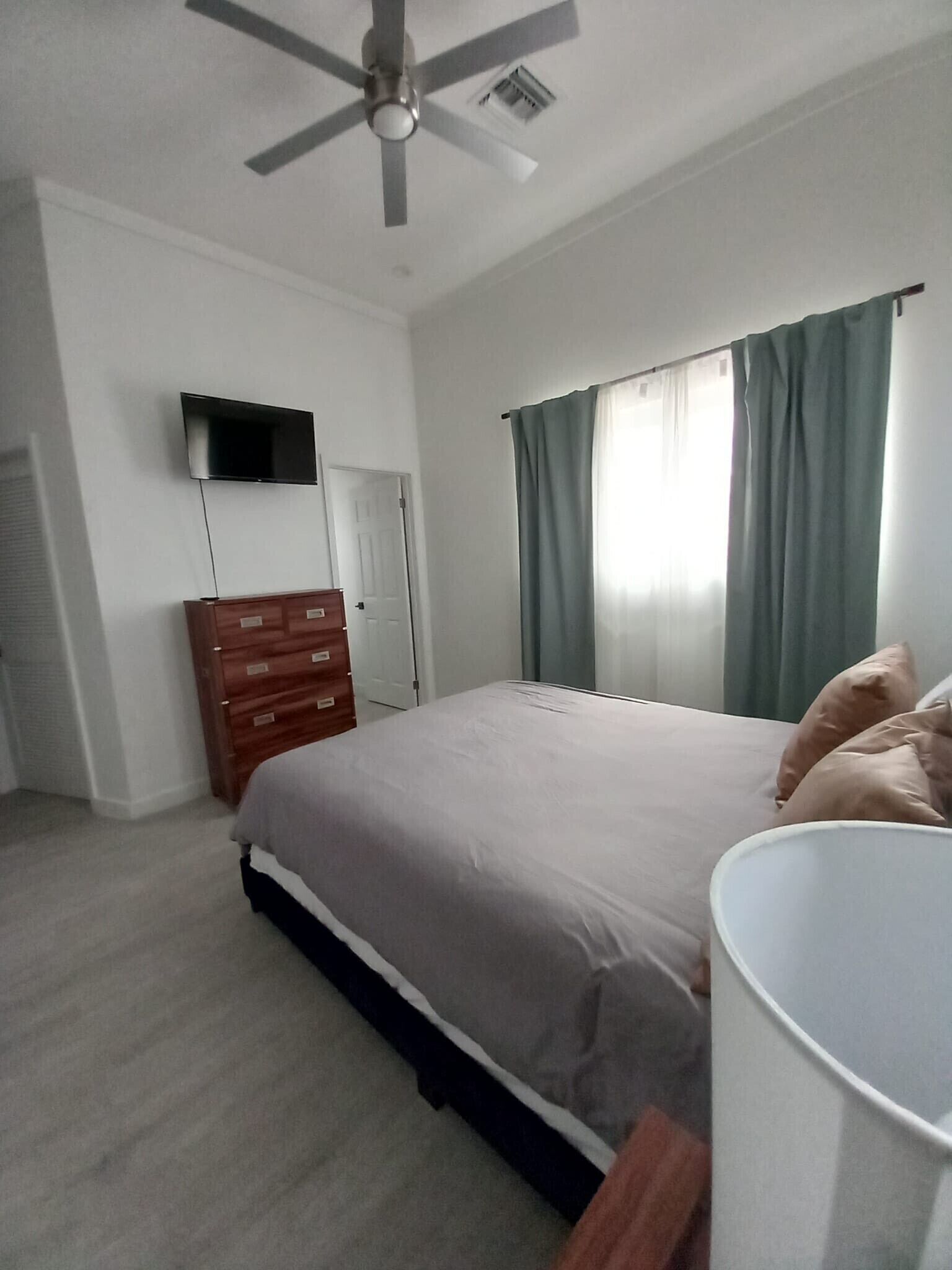 Una camera da letto con un letto, una cassettiera, una ventola a soffitto, un televisore fissato al muro e una finestra con tende.