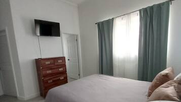 Una camera da letto con un letto, una cassettiera, una ventola a soffitto, un televisore fissato al muro e una finestra con tende.
