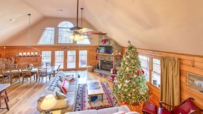 Een gezellig interieur van een chalet met een eethoek, een open haard, een kerstboom en een groot raam met uitzicht op een terras.