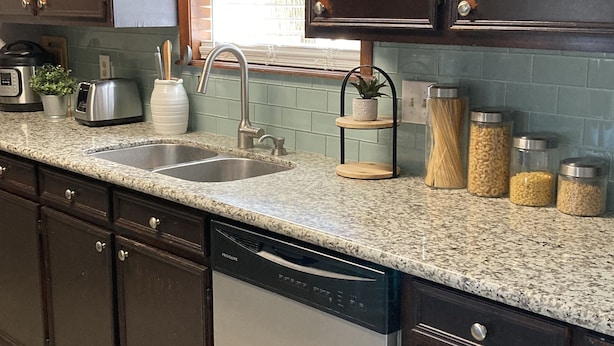 A kitchen with dark wooden cabinets, a granite countertop, a dishwasher, a sink, and a window with a blue curtain.
