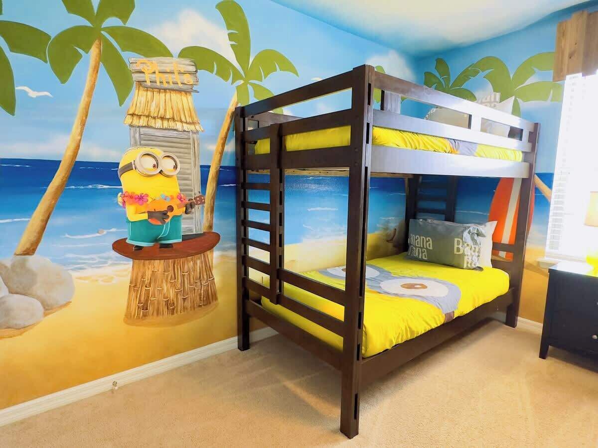 Una cama litera con un mural de playa tropical, un personaje de Minion y una mesita con un bolso.
