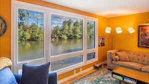 Ein Zimmer mit großen Fenstern, die auf einen See blicken, einem Sofa, einer Lampe und einem kleinen Tisch.