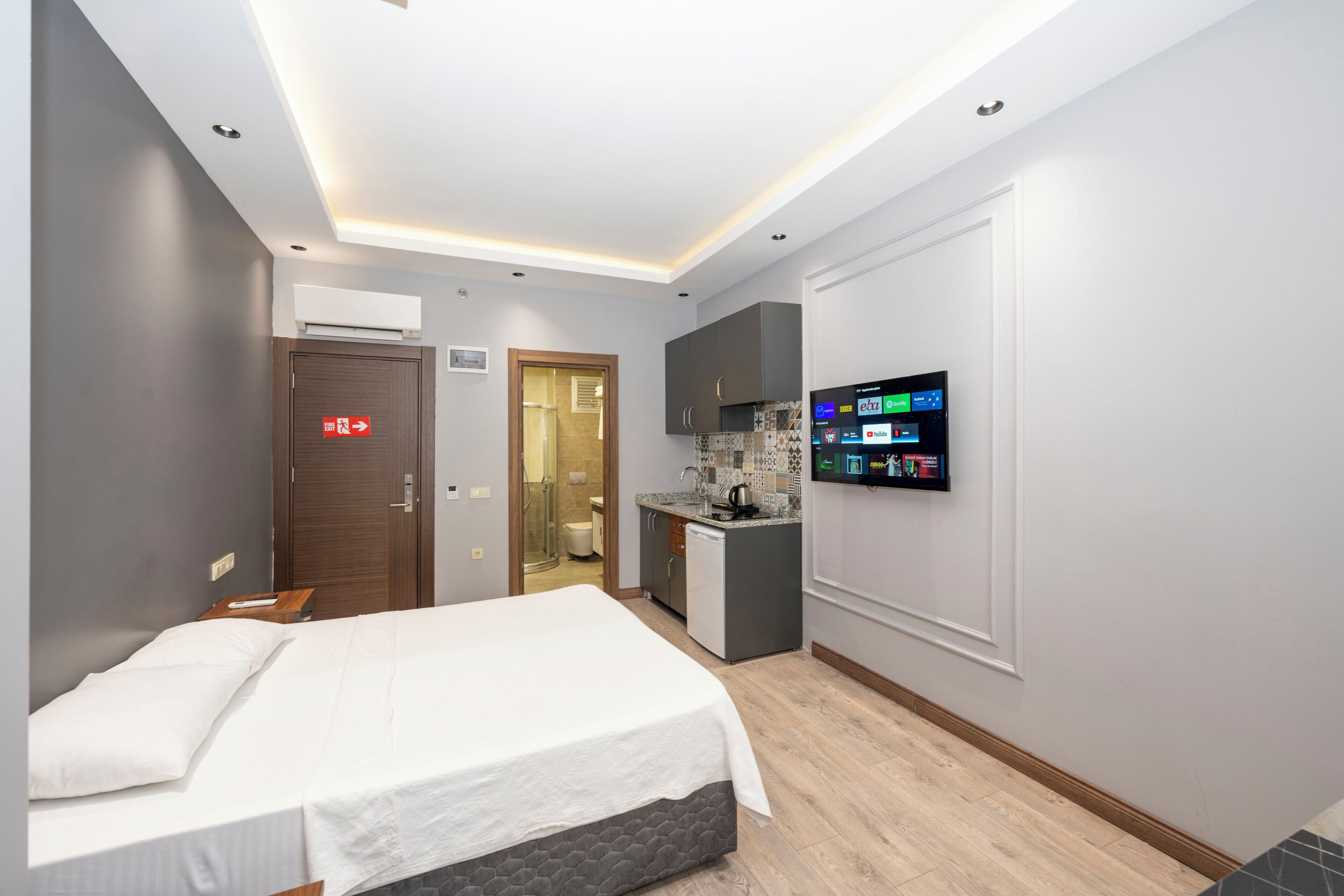 A modern hotel room with a large bed, a small kitchenette, and a mounted TV displaying various channels.