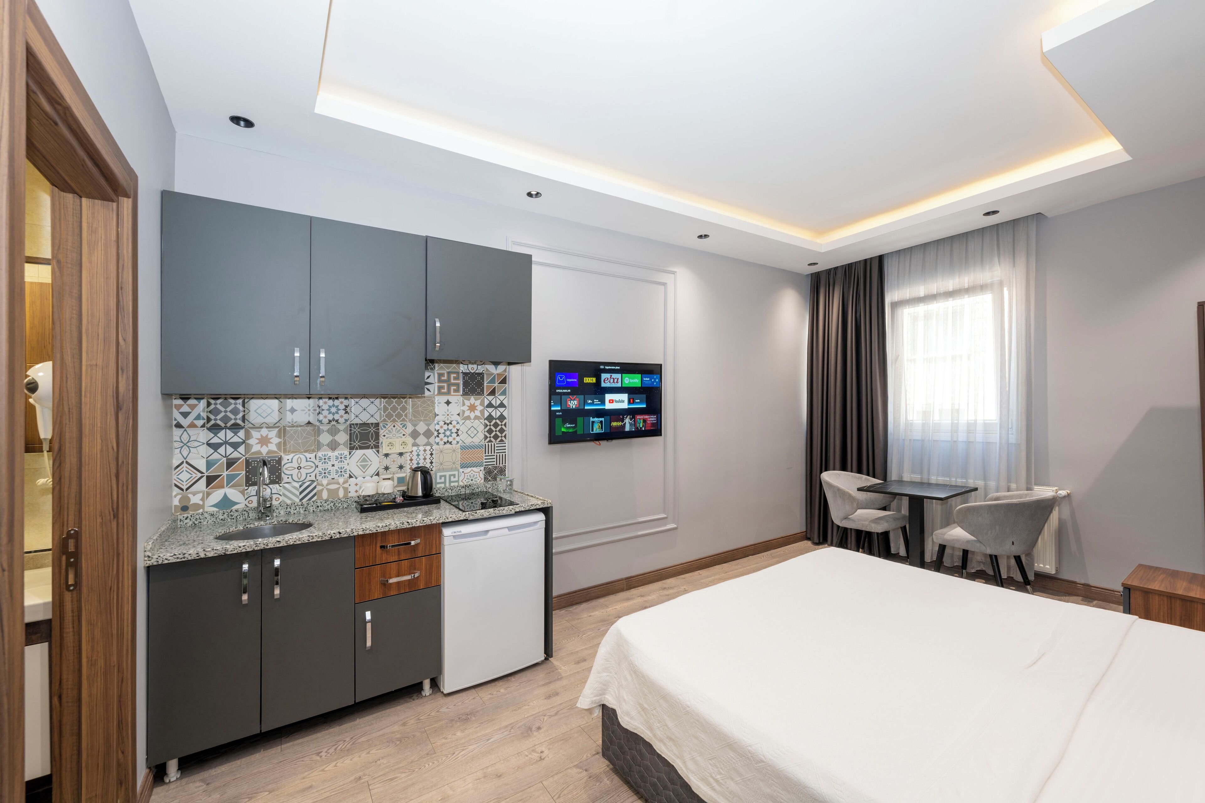 A modern hotel room with a kitchenette, a flat-screen TV, a bed, and a seating area with a table and chairs.