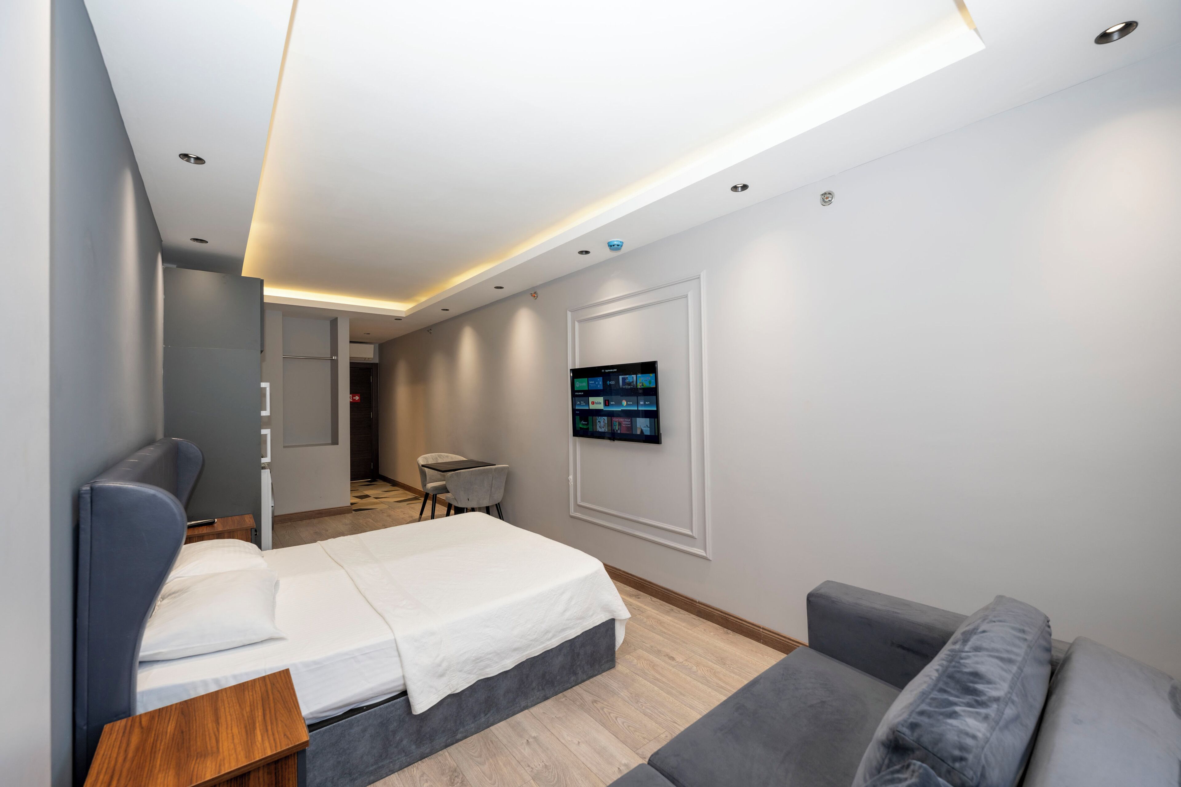 A modern hotel room with a bed, a grey sofa, a TV mounted on the wall, and a small desk.