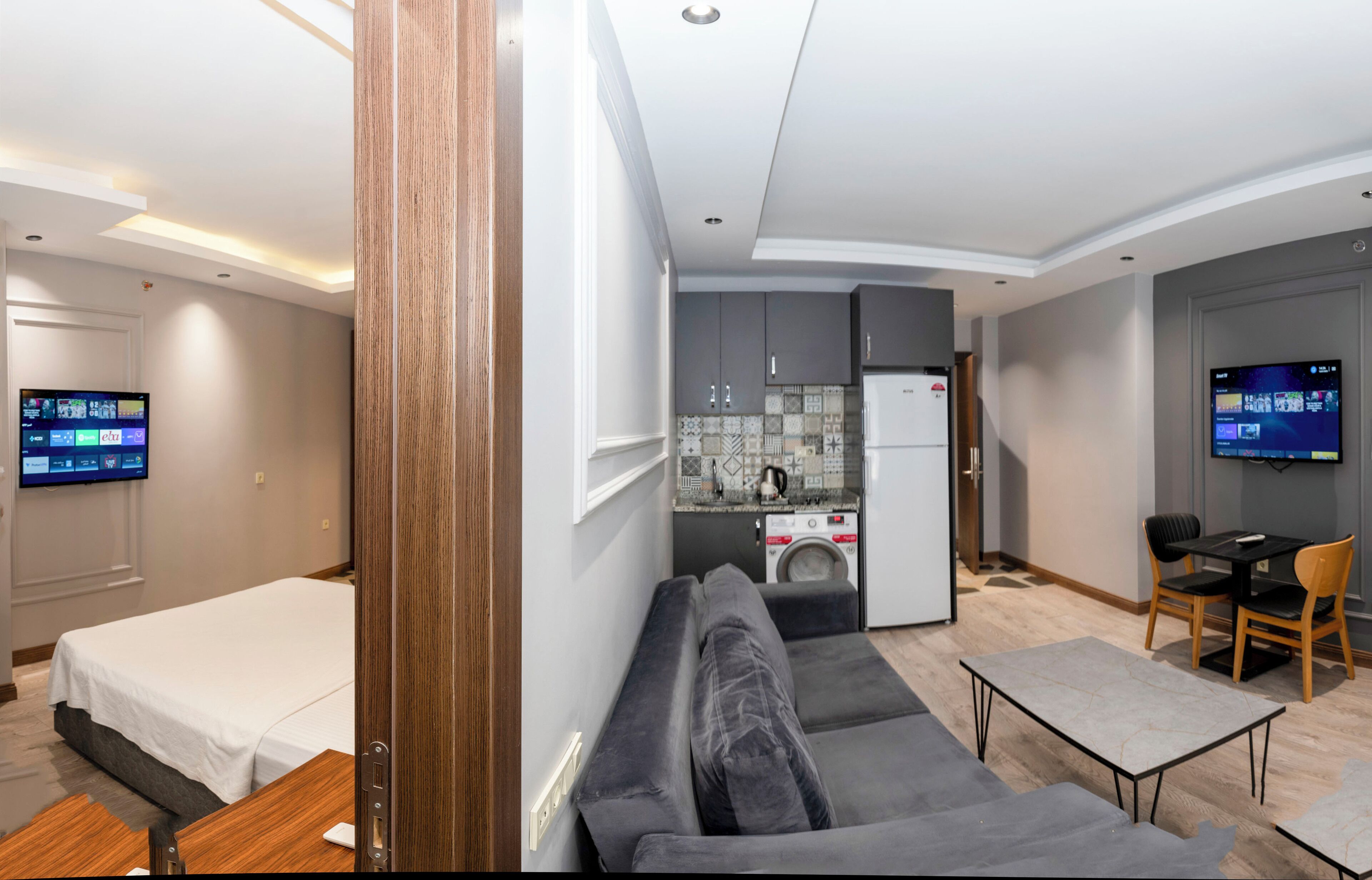 A modern hotel room with a sofa, a small dining table, a TV, and a kitchenette.