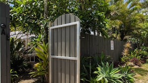A gated entrance with a metal gate and a wooden fence, surrounded by lush tropical plants and a stone pathway. - Alcorn Garden-Dog friendly 2 mins walk to beach (Suffolk Park)