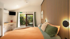A bedroom with a large bed, a TV mounted on the wall, a desk with a chair, and a view of the garden through an open door. - Alcorn Garden-Dog friendly 2 mins walk to beach (Suffolk Park)