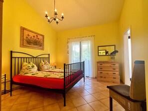 Ein Schlafzimmer mit einem Bett, einem Schminktisch, einem Sessel, einer Deckenleuchte und einem Fenster mit Vorhängen.