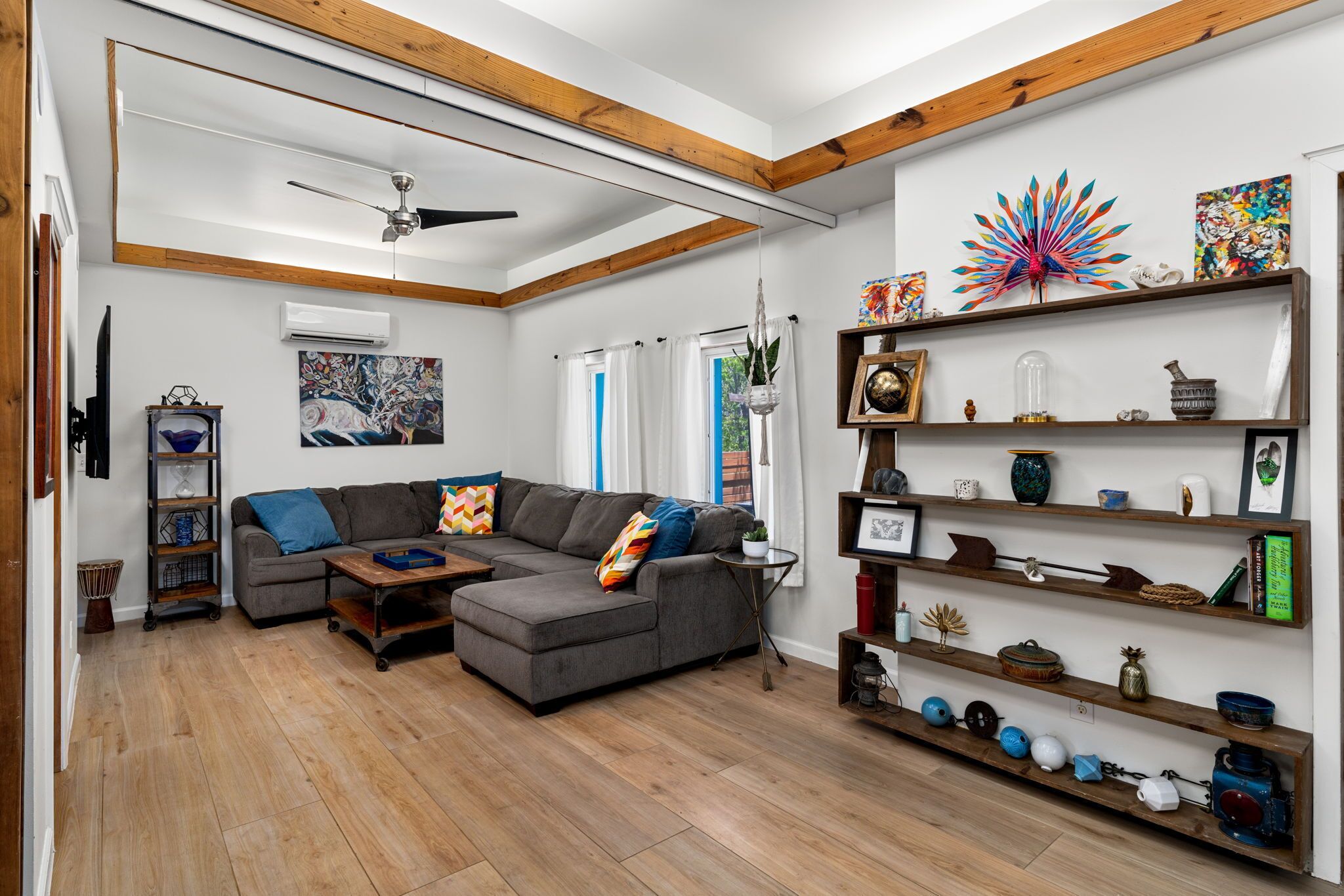 Een moderne woonkamer met een hoekbank, houten balken, een plafondventilator en diverse decoratieve objecten op planken.
