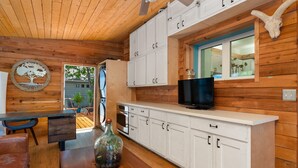 A wooden interior with a kitchen area featuring white cabinets, a black television, and a glass bottle on a wooden table. - Boho-Ranch Retreat in Hip East Austin w Hot Tub and Cowgirl pool. (Austin)