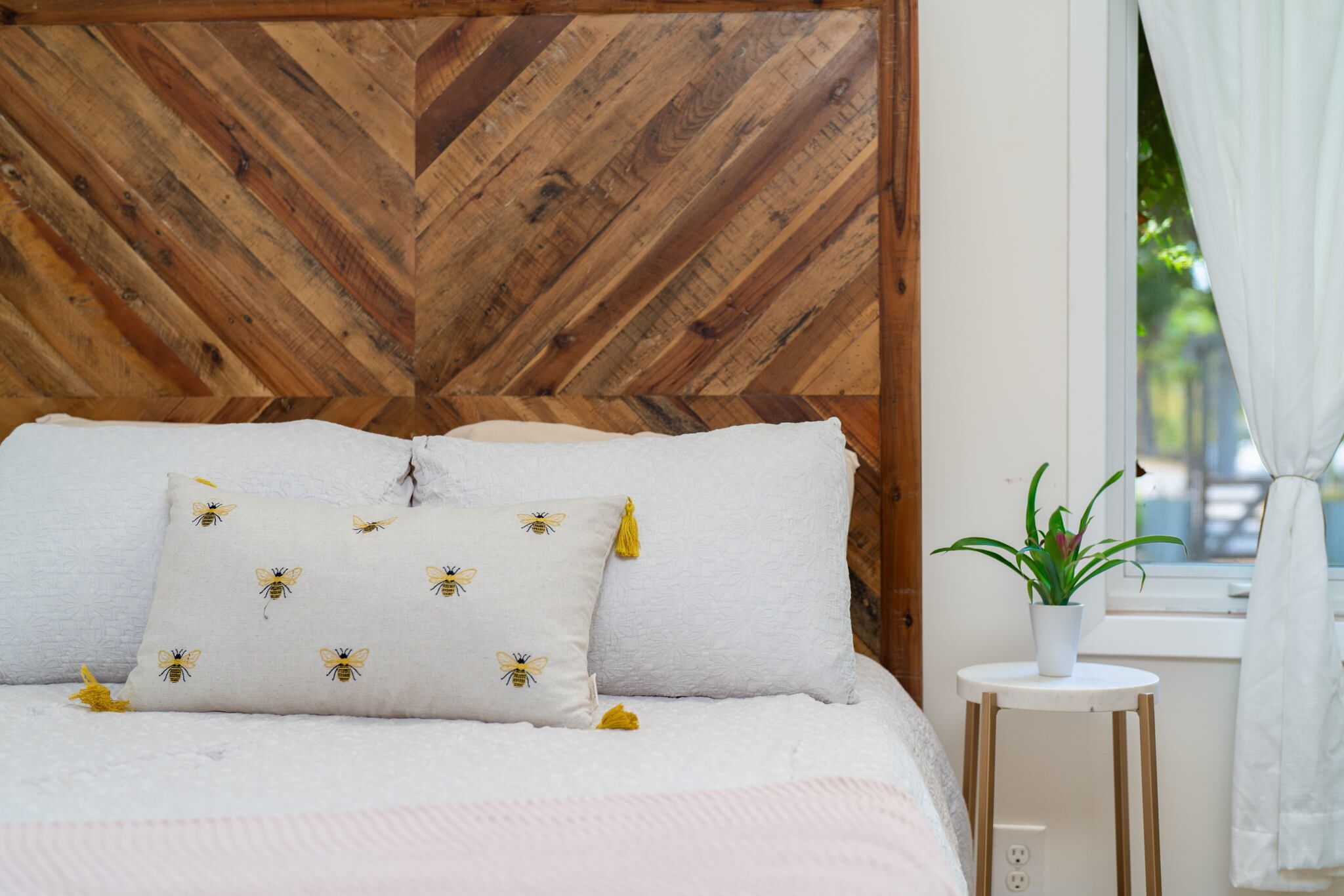 Una cama con ropa de cama blanca y almohadas con estampado de abejas, un cabecero de madera, una mesita de noche blanca con una planta y una ventana con cortinas blancas.