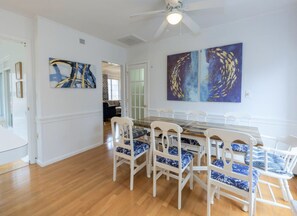 A dining area with a glass-top table and white chairs with blue patterned cushions. There are abstract paintings on the walls and a ceiling fan above.