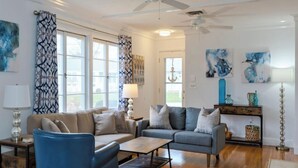 Ein geräumiges Wohnzimmer mit einer Couch, einem Sessel und einem Couchtisch. Zwei Fenster mit Vorhängen, ein Deckenventilator und Kunstwerke an den Wänden.