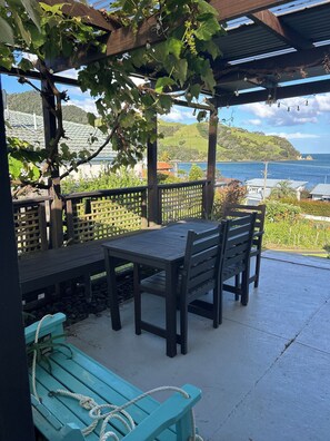 A covered outdoor seating area with a table and chairs, a bench, and a view of the sea and hills. - Simpson's Beach - Fantastic View - Beach Home (Whitianga)