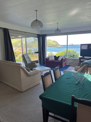A spacious living room with a large window offering a scenic view of the sea and hills. There is a beige sofa, a purple sofa, and a dining table with chairs. - Simpson's Beach - Fantastic View - Beach Home (Whitianga)