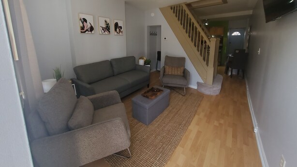 Un salon avec un canapé gris, un petit pouf et un fauteuil. On y trouve un escalier avec une rampe en bois qui mène à une autre pièce.