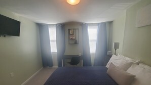 Une chambre avec un lit, un bureau et une chaise. Il y a deux fenêtres avec des rideaux, un luminaire au plafond et un téléviseur fixé au mur.